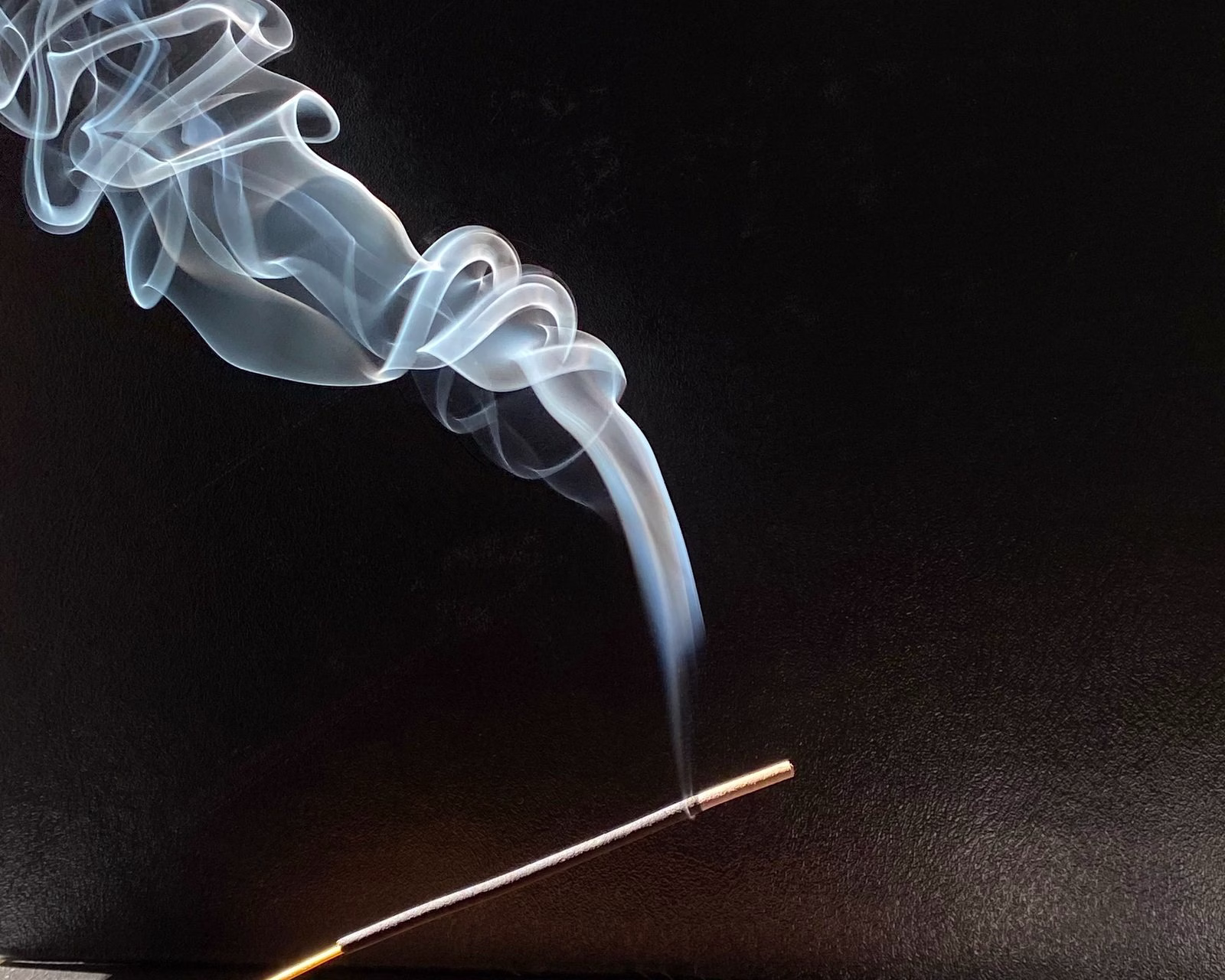 Burning incense stick on a wooden holder with a curl of pale blue smoke rising against a black backdrop