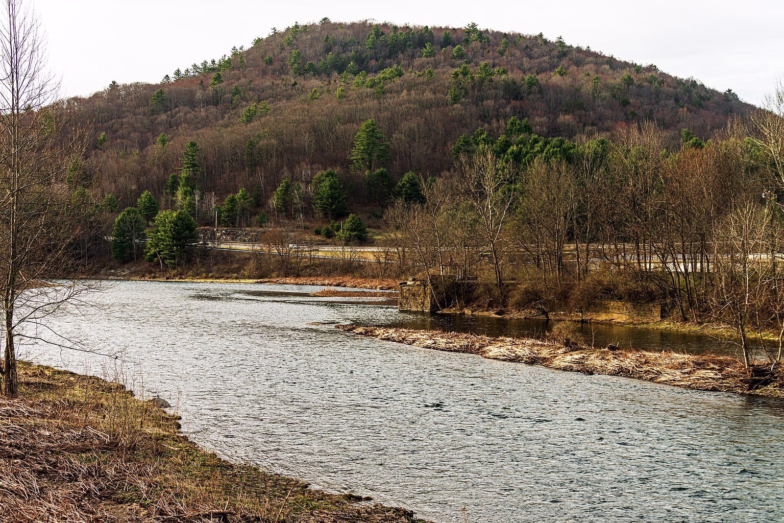 A wide river bend below a wooded hill, with a gazebo on a stone wall at the water.