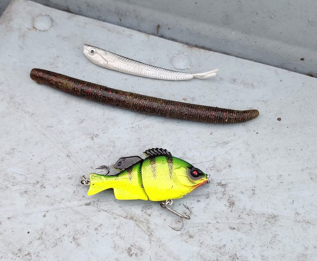 Three lures on the gray floor of the boat: a white soft jerkbait, a dark Yamamoto Senko stick worm with red flake, and a chartreuse jointed sunfish swimbait.