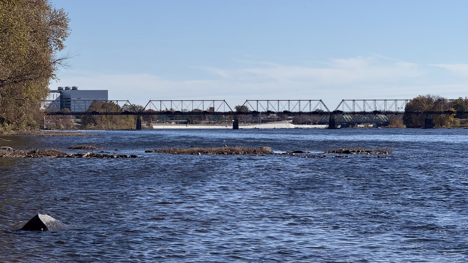 Rippled blue water over rocky bars, with a long truss bridge in the distance.