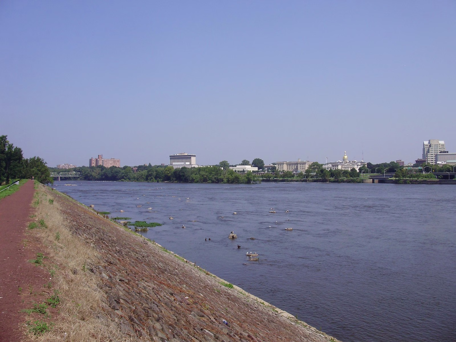 A rock-lined bank and wide riffled water, with a city skyline and a gold dome across the river.