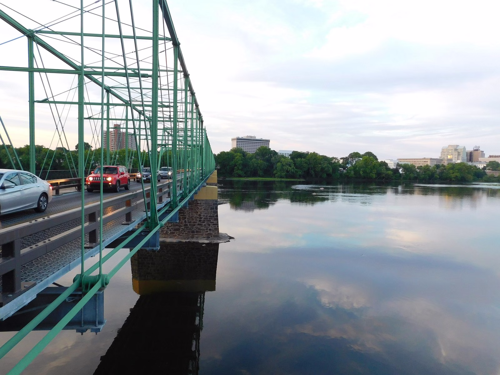 A green truss bridge on stone piers over still water reflecting the sky, with city buildings behind.
