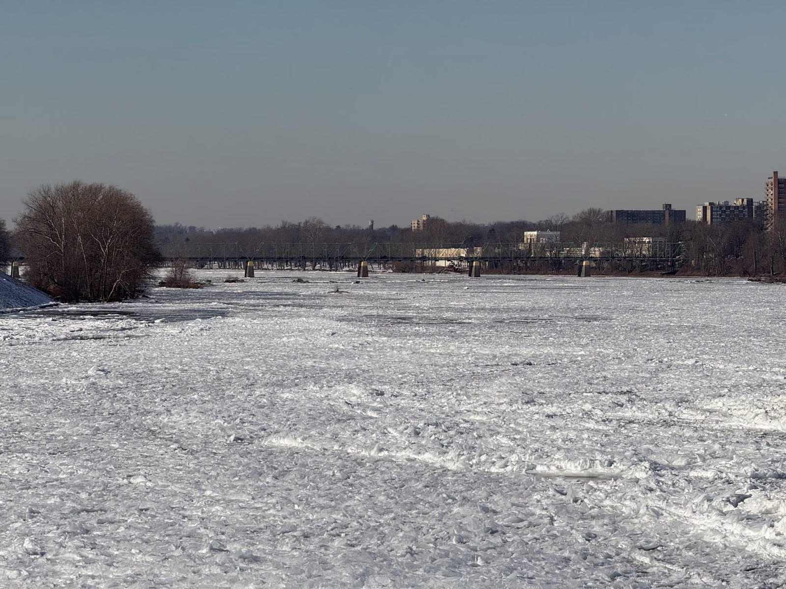A river covered in broken white ice, with a bridge and buildings in the distance.