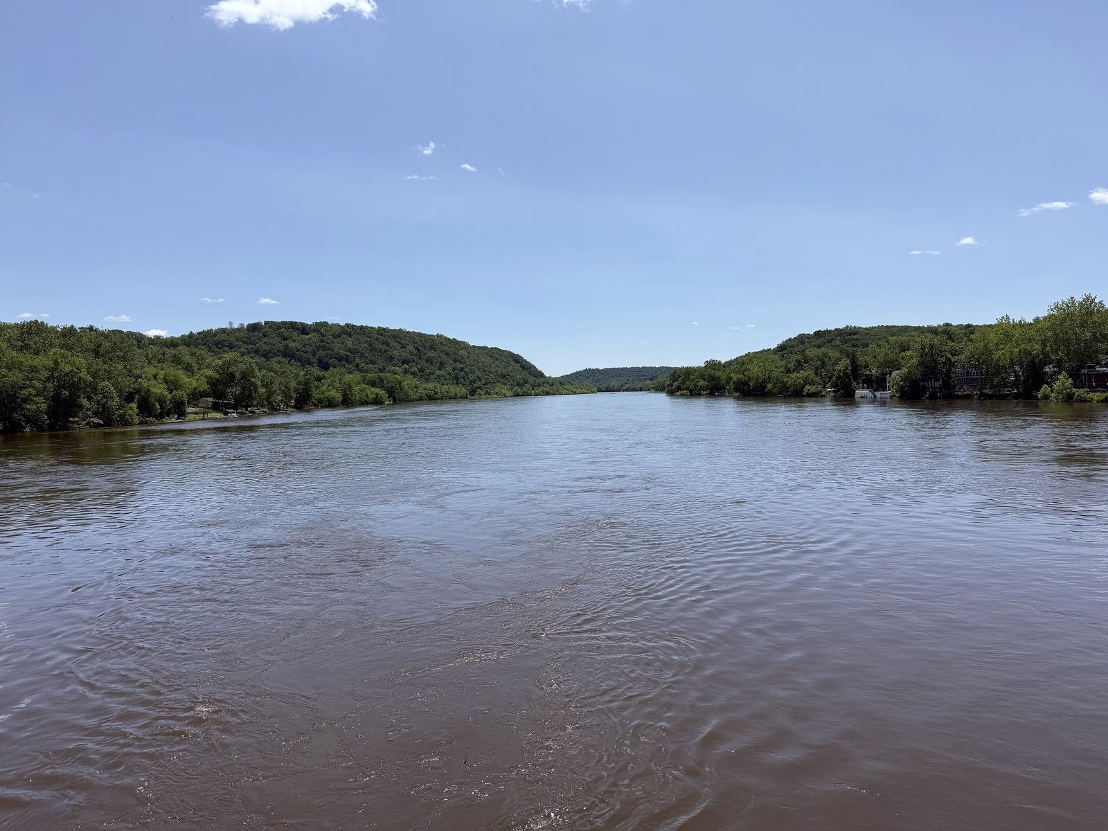 A wide brown river running between green wooded hills under a blue sky.