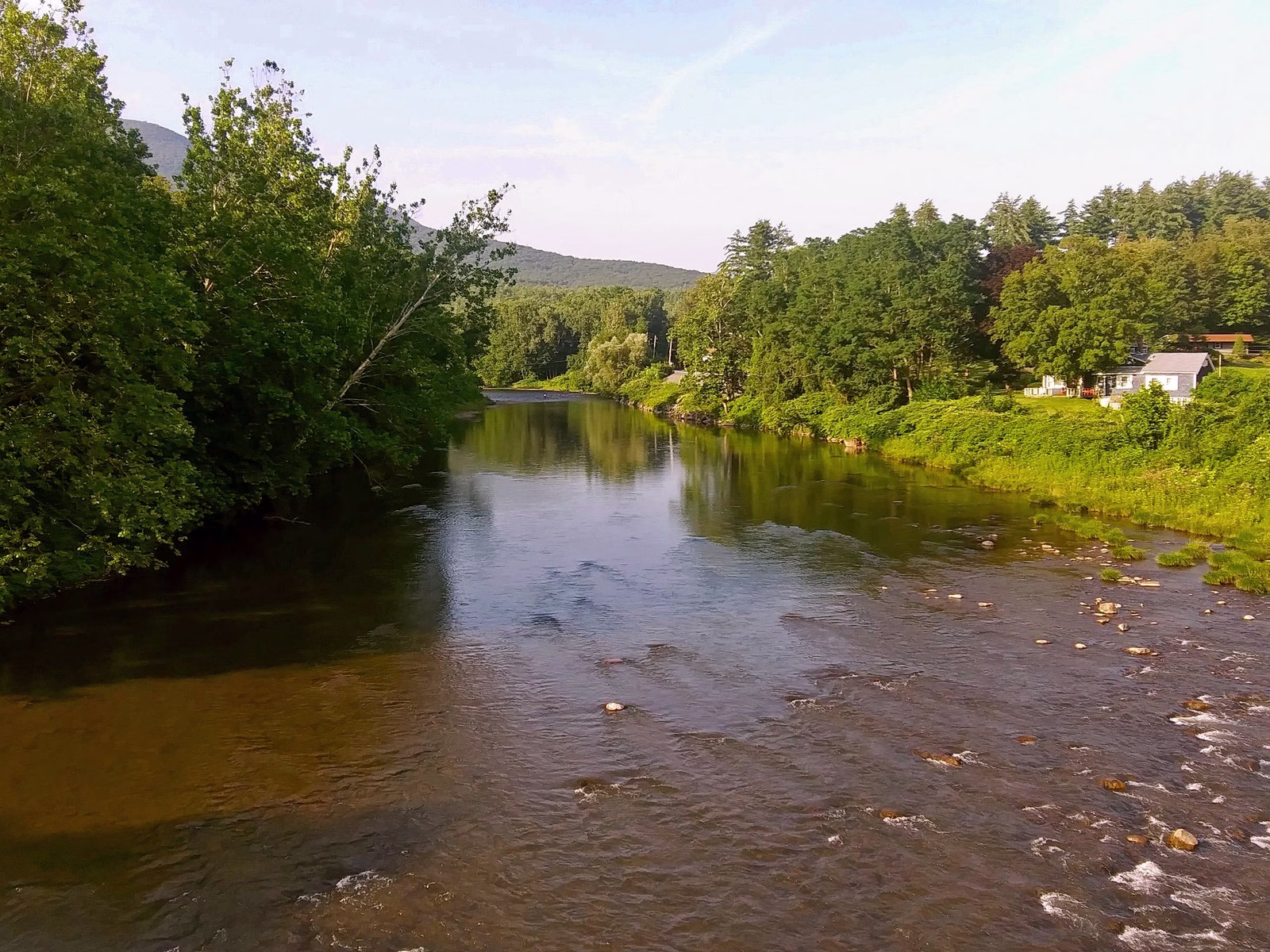 A calm pool sliding into riffles, with houses on the right bank and a mountain behind.