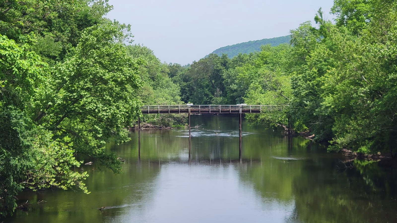 A wide, calm green river with a footbridge in the distance and a hill behind.