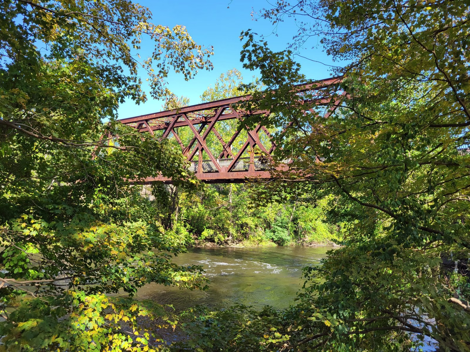 A red steel bridge seen through autumn leaves above a riffled river.