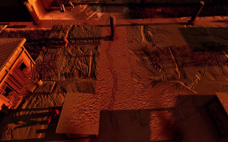 A town street seen from above at dusk in warm orange light, with square dirt plots, a lone stone column, a house with a tiled roof on the left and a few small figures.