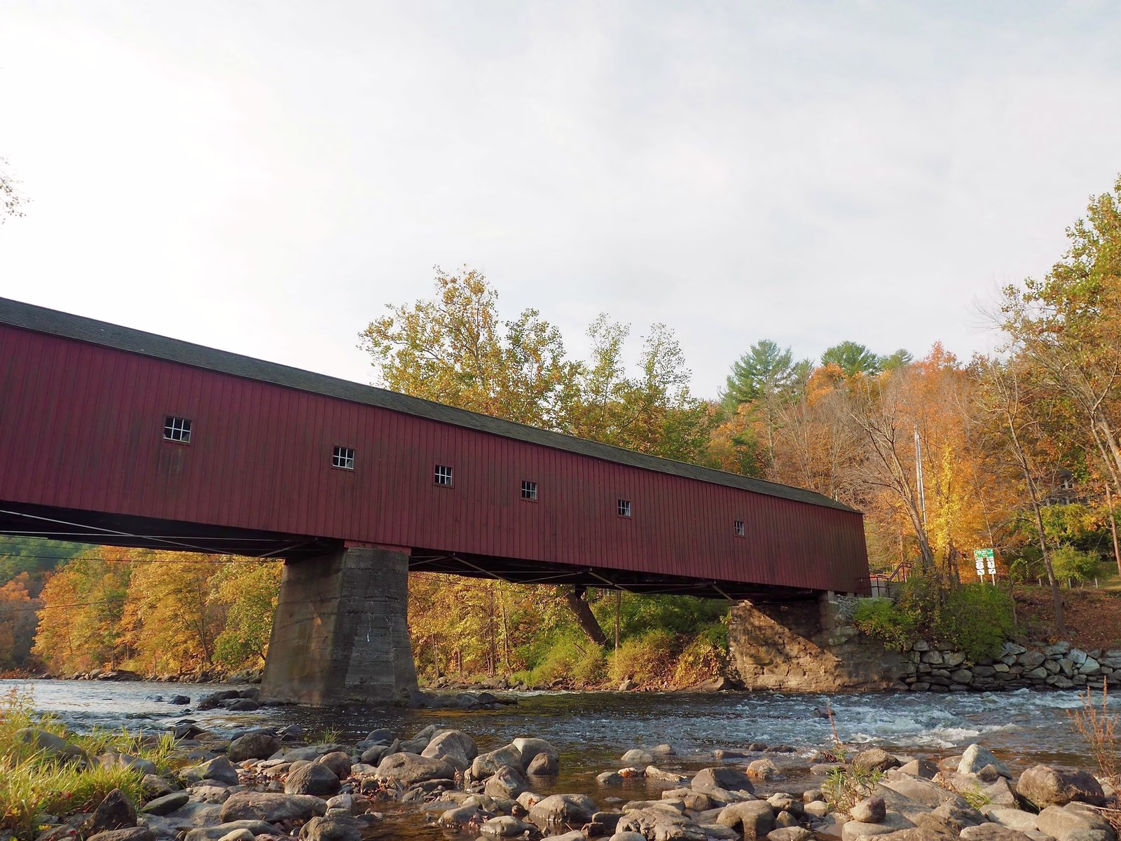 A long red covered bridge over a rocky riffle, with orange and gold trees behind.