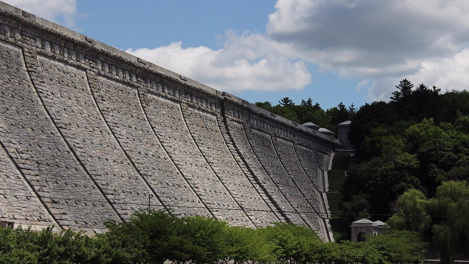 The tall curved stone face of a dam under a blue sky with white clouds, with a row of green trees at its foot.