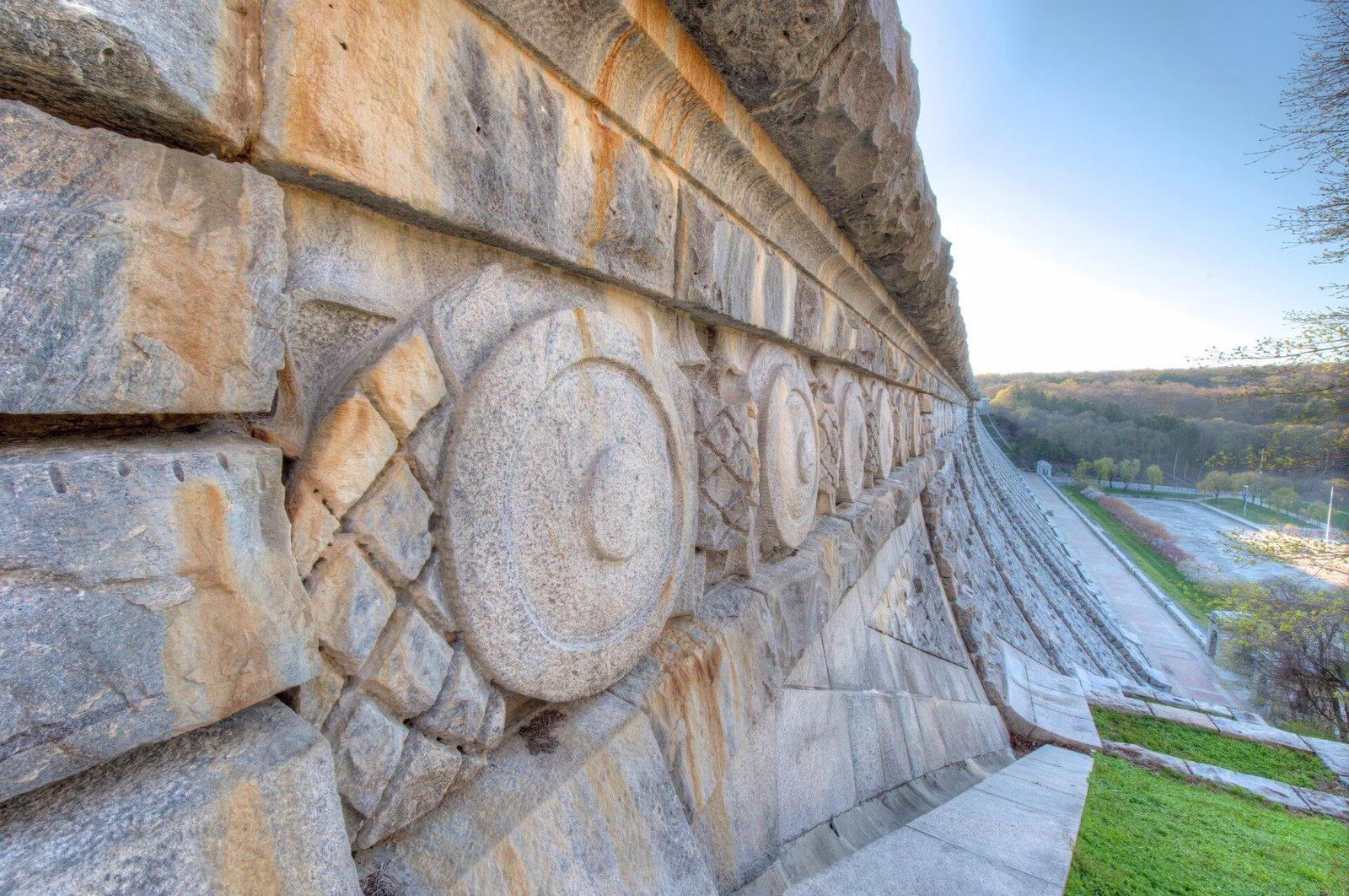 A close view of big carved stone discs along the crest of a dam, with a plaza far below.