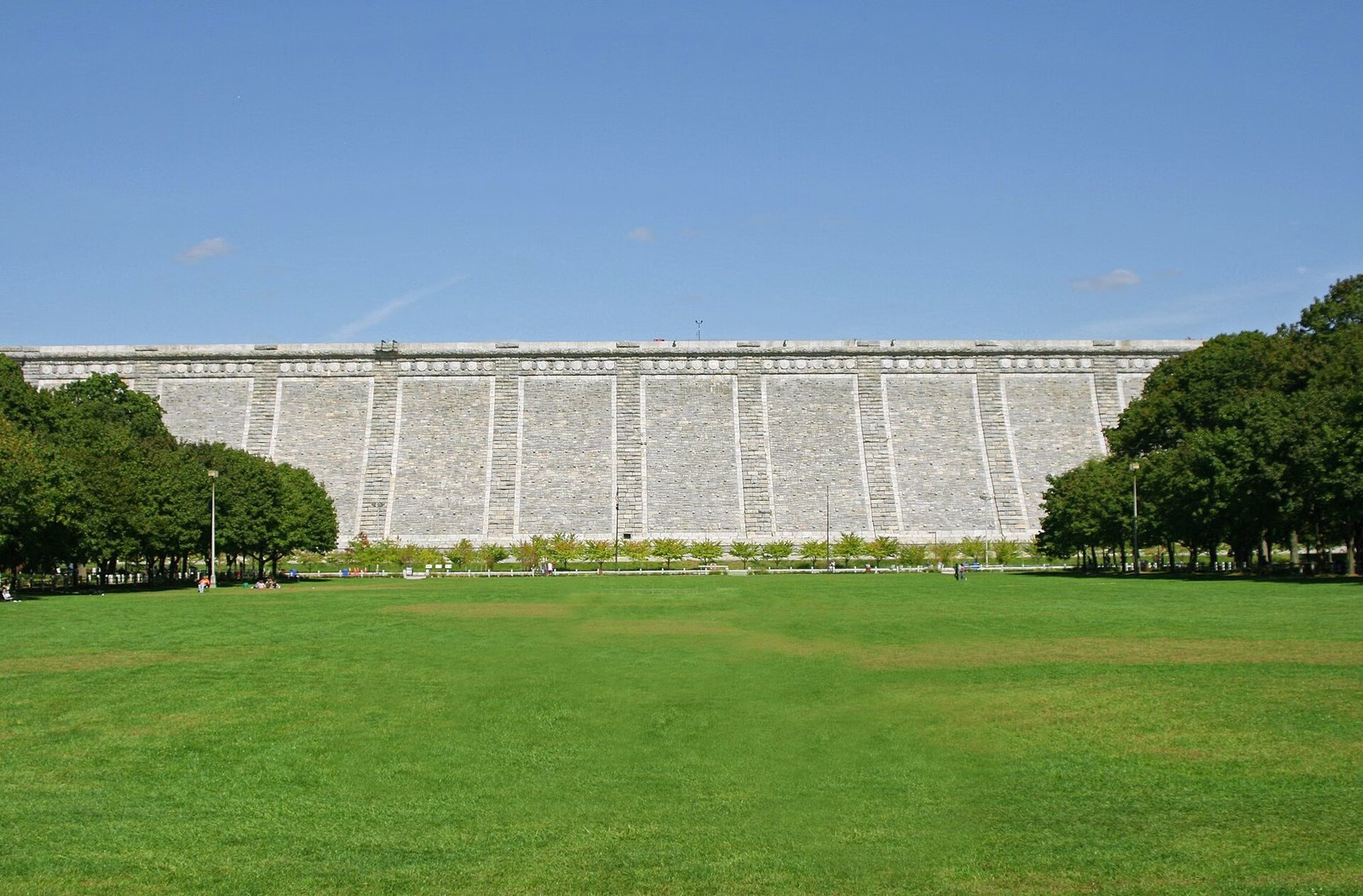 A long, flat stone dam face across the end of a wide green lawn under a blue sky.