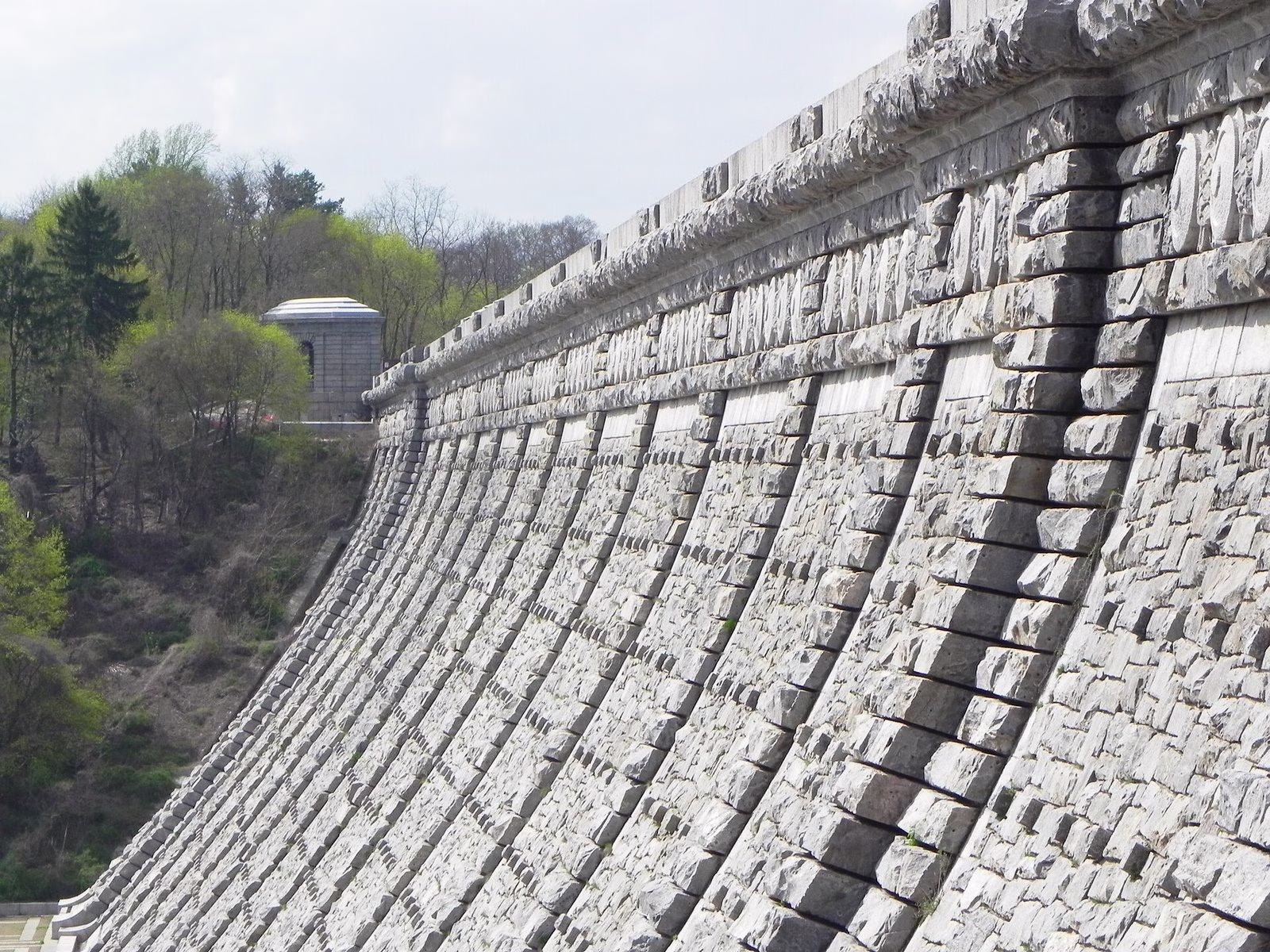 Rows of stepped grey stone curving up the face of a dam, with a small stone pavilion and spring trees behind.
