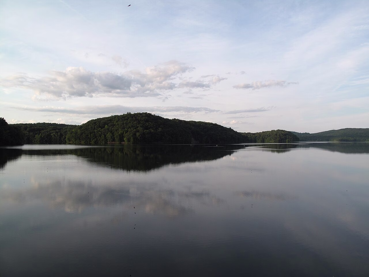 Still water reflecting a pale evening sky, with a dark wooded hill across the reservoir.