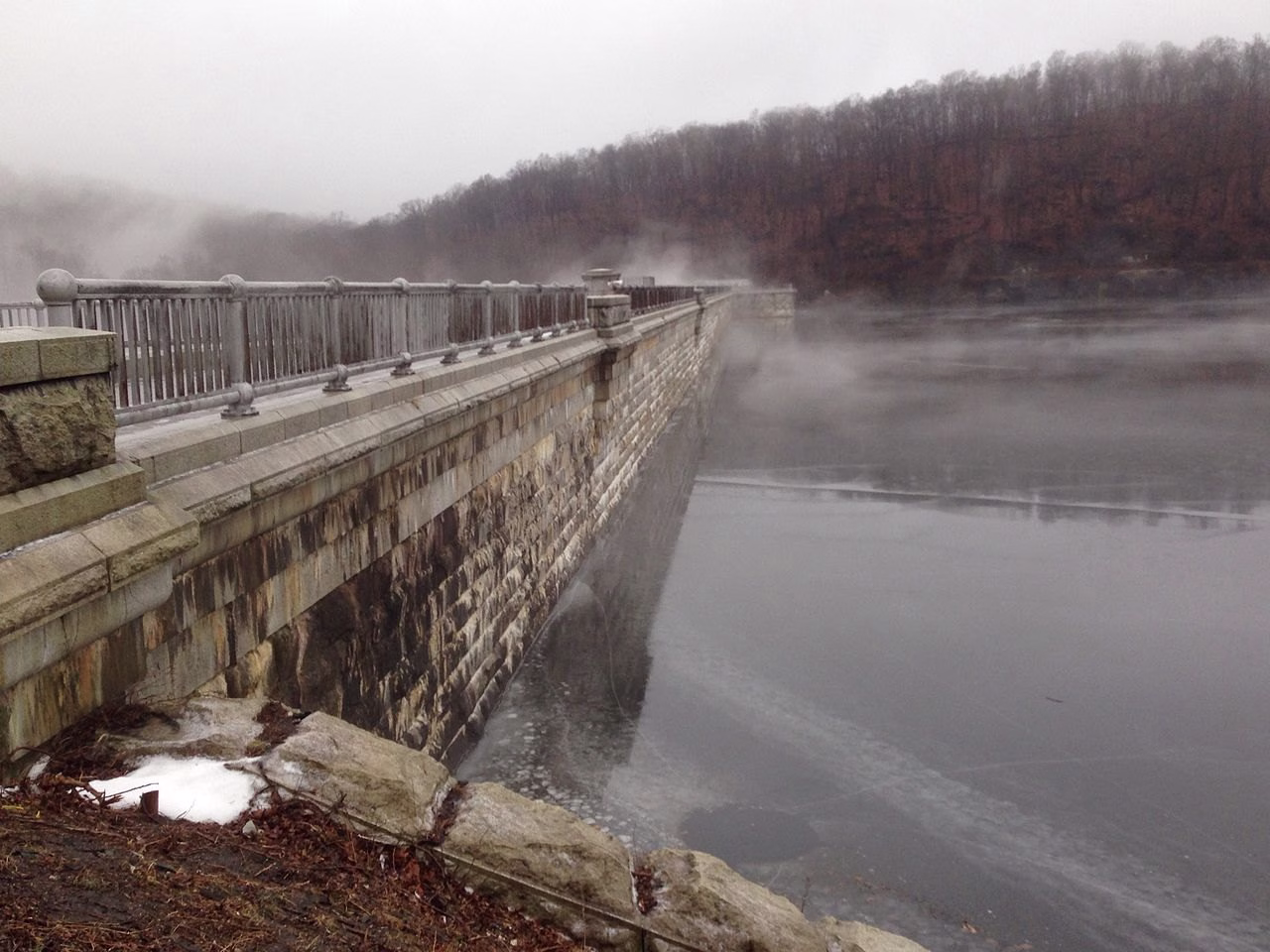 A long stone dam wall with a railing running into mist over calm grey water, with bare hills behind.