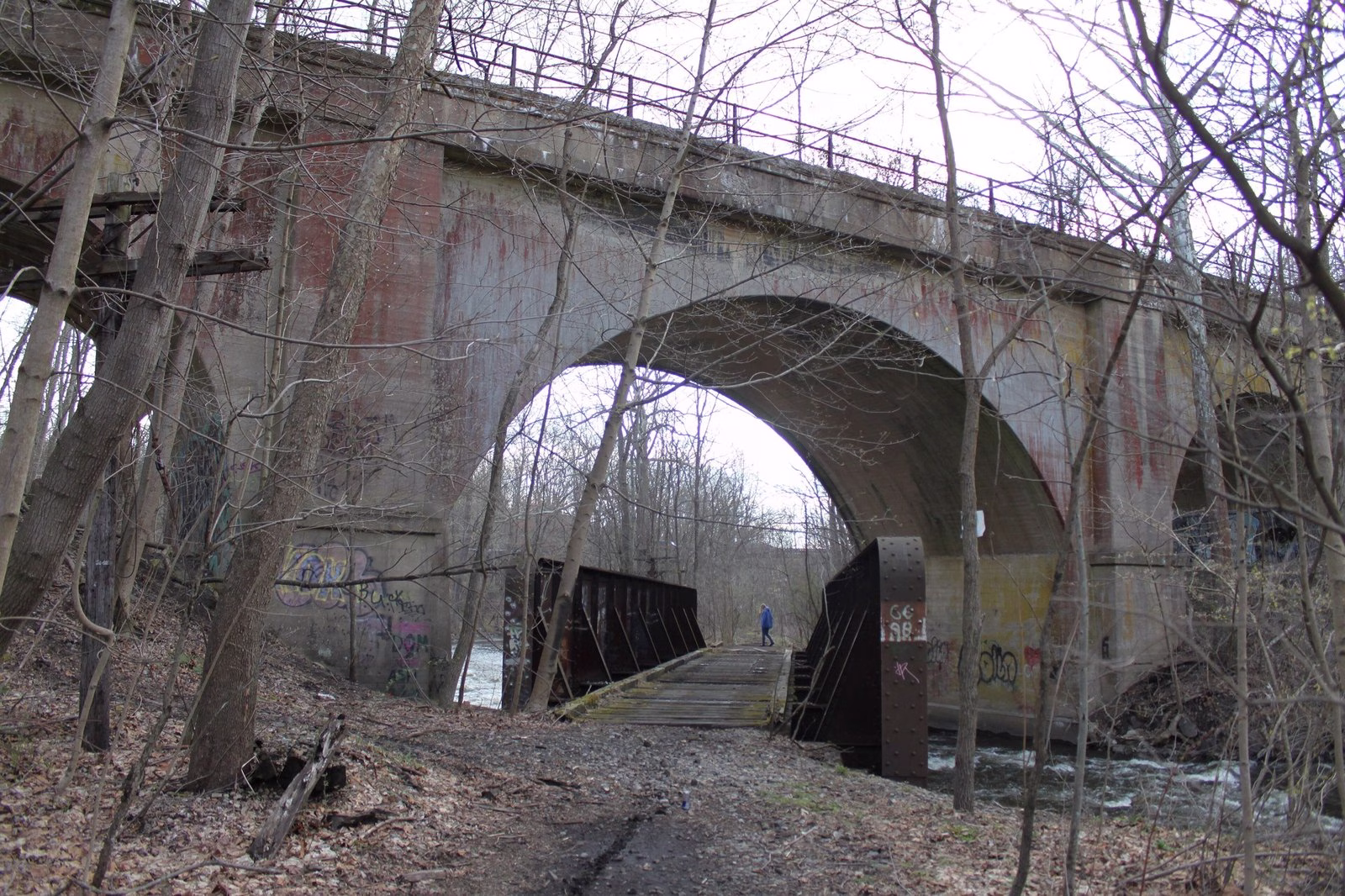 A tall concrete arch bridge above a small river and a footbridge, among bare spring trees.