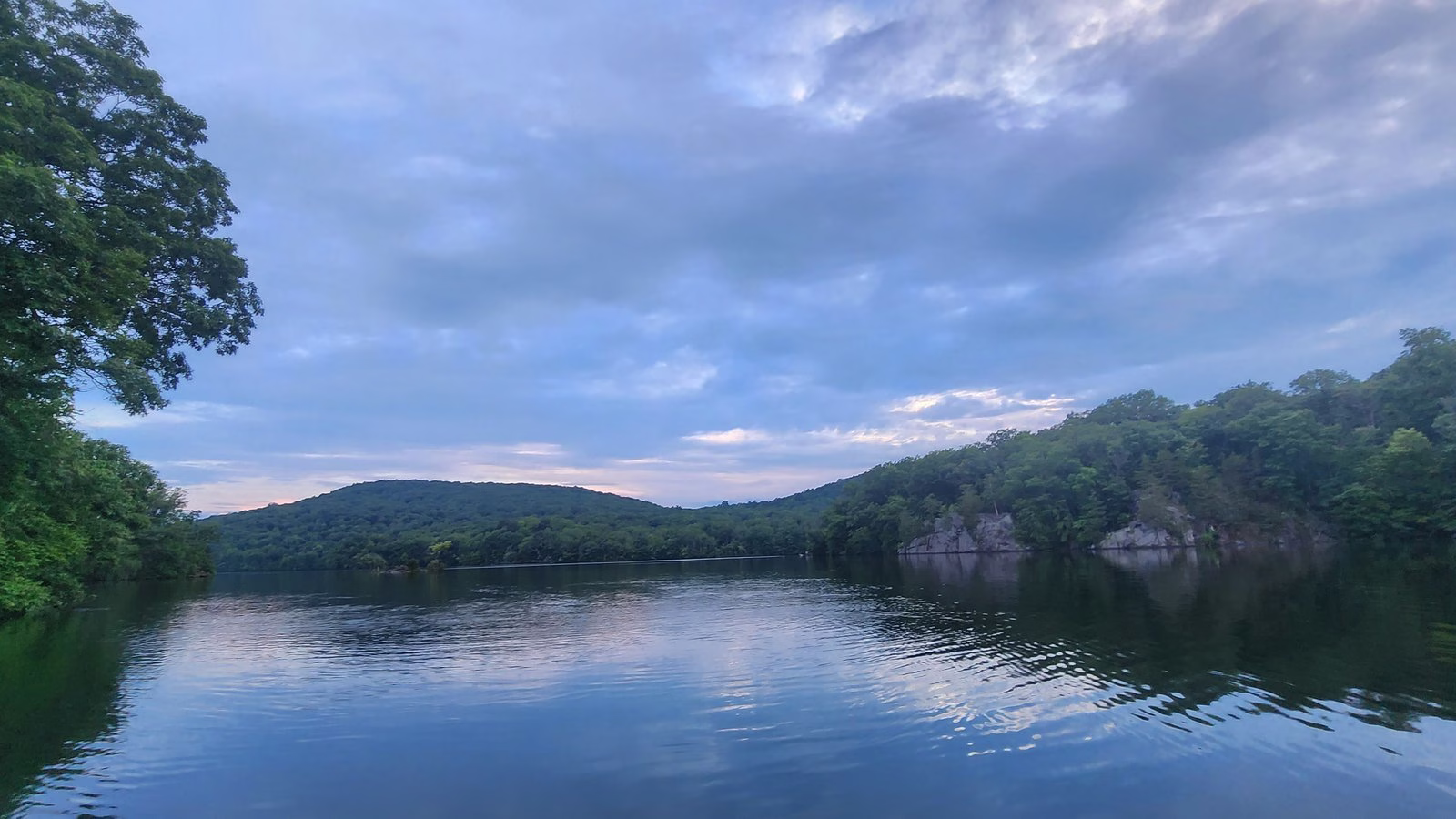 A wide, calm reservoir under a soft blue evening sky, with wooded hills and grey rock ledges along the shore.