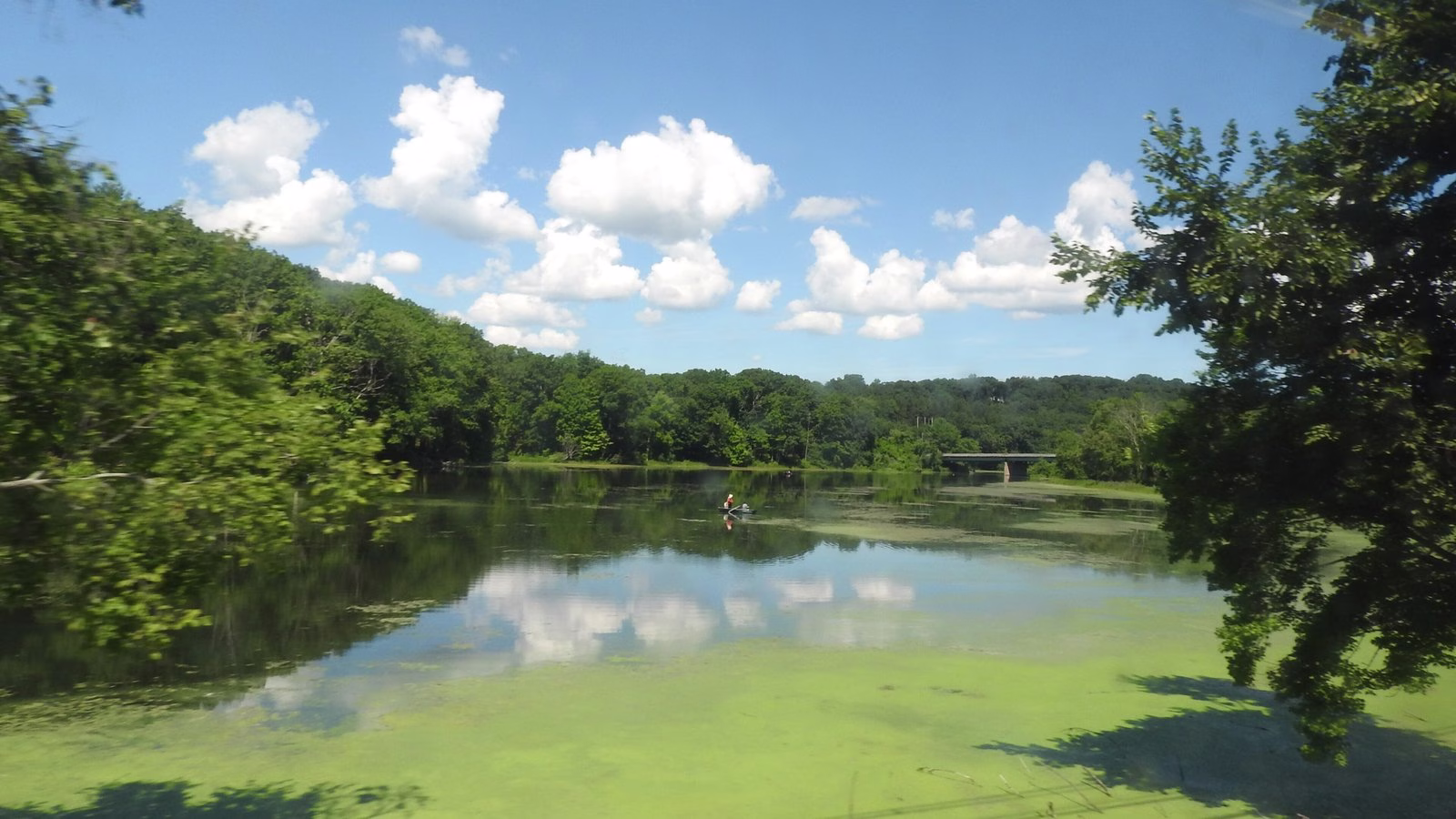 A calm river reflecting puffy clouds, with a small boat far out and a bridge beyond.