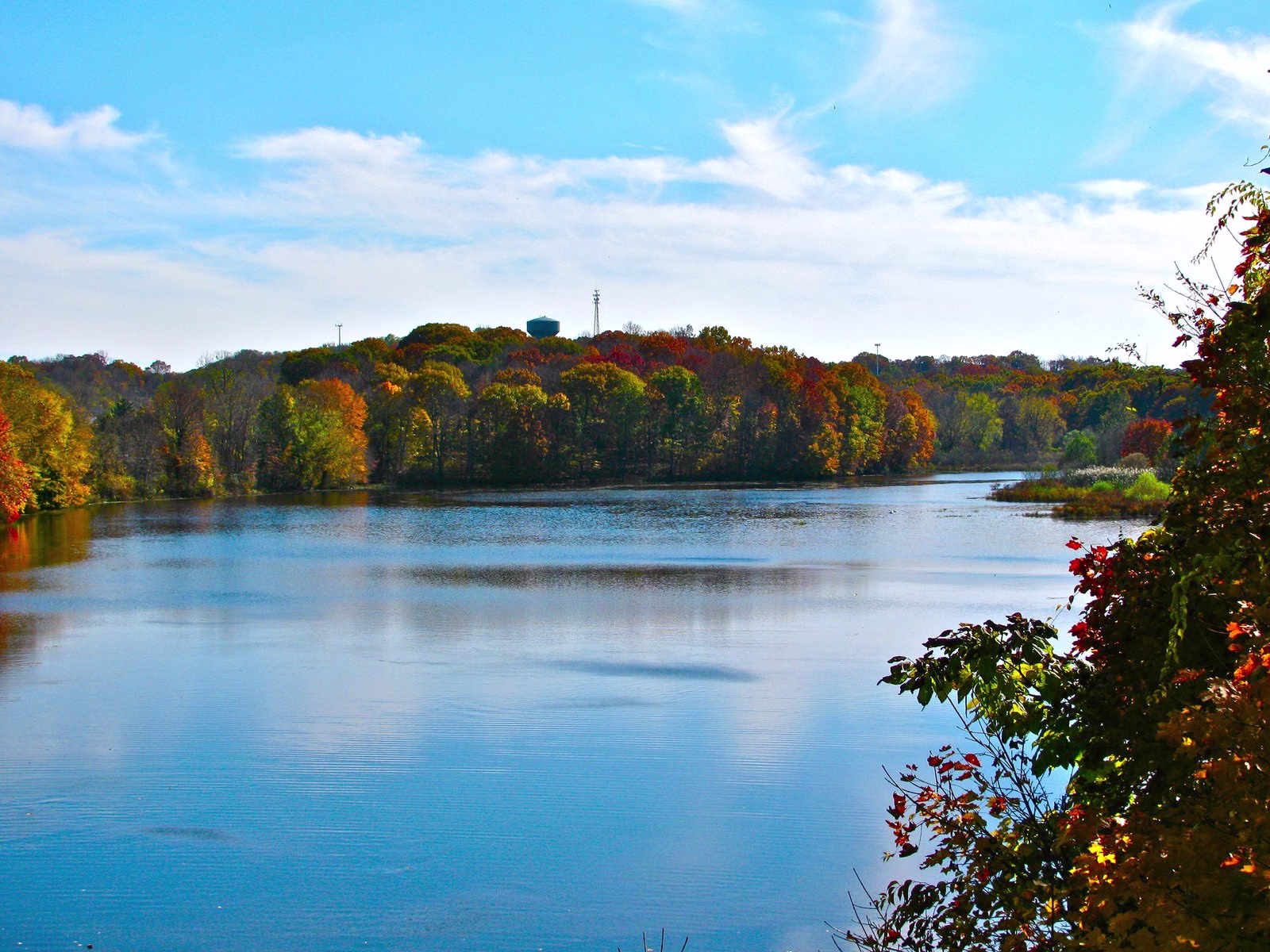 A calm blue reservoir under orange and red fall trees and a bright sky.